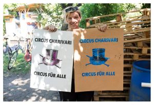 Foto von Friederike Charivari die zwei Poster in der Hand hällt. Auf dem Poster steht jeweils "Circus Charivari Circus für alle" und das Hut-Logo vom Circus Charivari